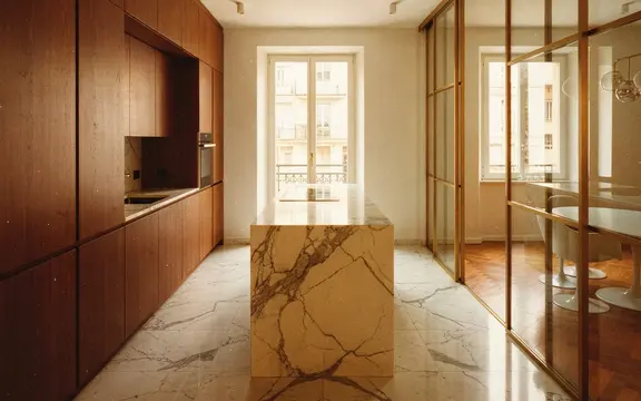 Cucina con isola in marmo Calacatta e boiserie in noce; parete vetrata con montanti in ottone verso la zona pranzo; luce naturale da finestra con affaccio su Milano
