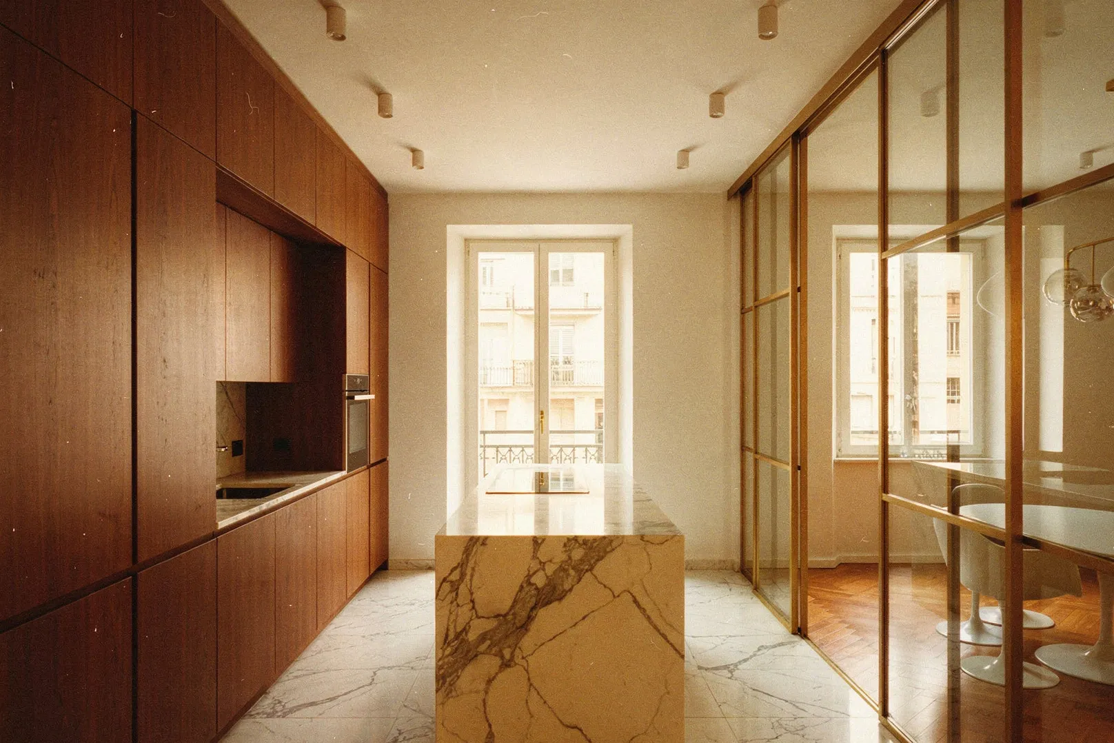 Cucina lineare con ante in noce scuro; isola in marmo Calacatta bianco venato; parete vetrata con montanti sottili in ottone verso la zona pranzo; finestre con affaccio su edifici milanesi