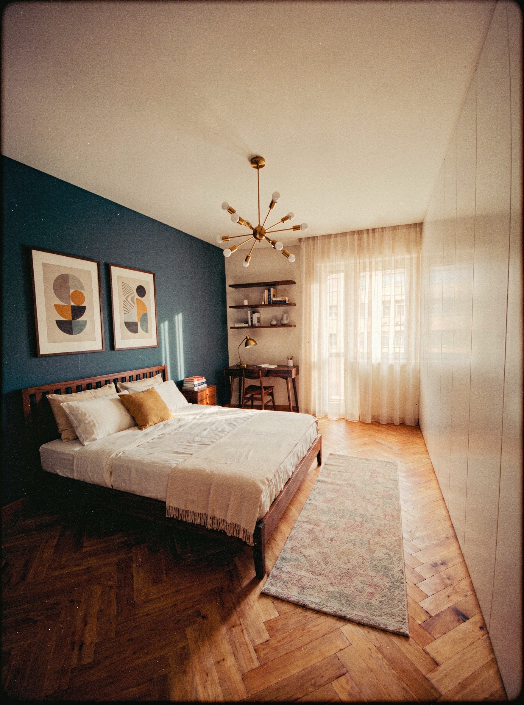 Seconda camera con parete blu petrolio; letto in legno; due stampe geometriche astratte; angolo studio con scrivania in noce; lampadario sputnik in ottone; armadio bianco a tutta altezza; tappeto geometrico blu e beige; pavimento a spina di pesce
